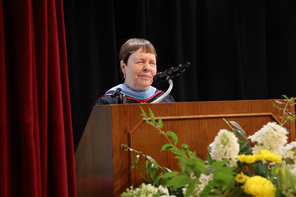 The commencement speaker for Alabama School for the Blind speaks into a microphone while standing behind a podium. She wears black ceremonial robes and a light blue hood.